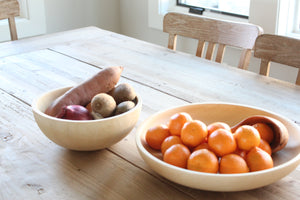 A mango wood plate filled with oranges next to a mango wood bowl with various fruits/vegetables